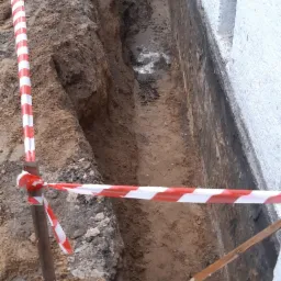 Wykop wzdłuż ściany budynku, zabezpieczony taśmą ostrzegawczą, z widocznymi fragmentami rur kanalizacyjnych i instalacji.
