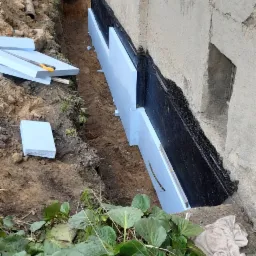 Ocieplenie fundamentów budynku styropianem w wykopie, widoczne warstwy izolacji bitumicznej i fragmenty styropianu na ziemi.