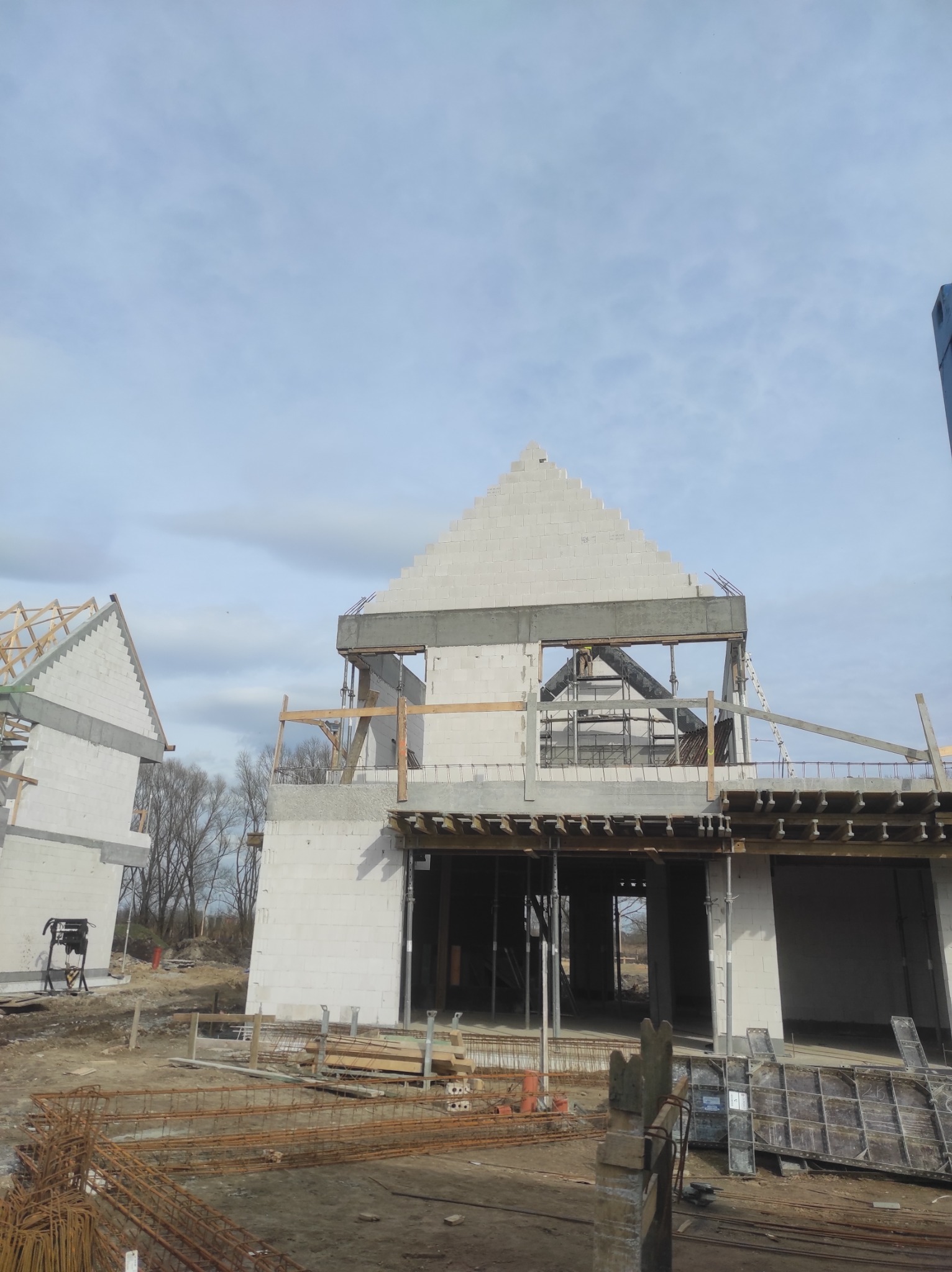 Budowa domu z białych bloczków: widok na surowy stan z drewnianym szalunkiem i zbrojeniami, w tle drugi budynek w trakcie wznoszenia pod pochmurnym niebem.