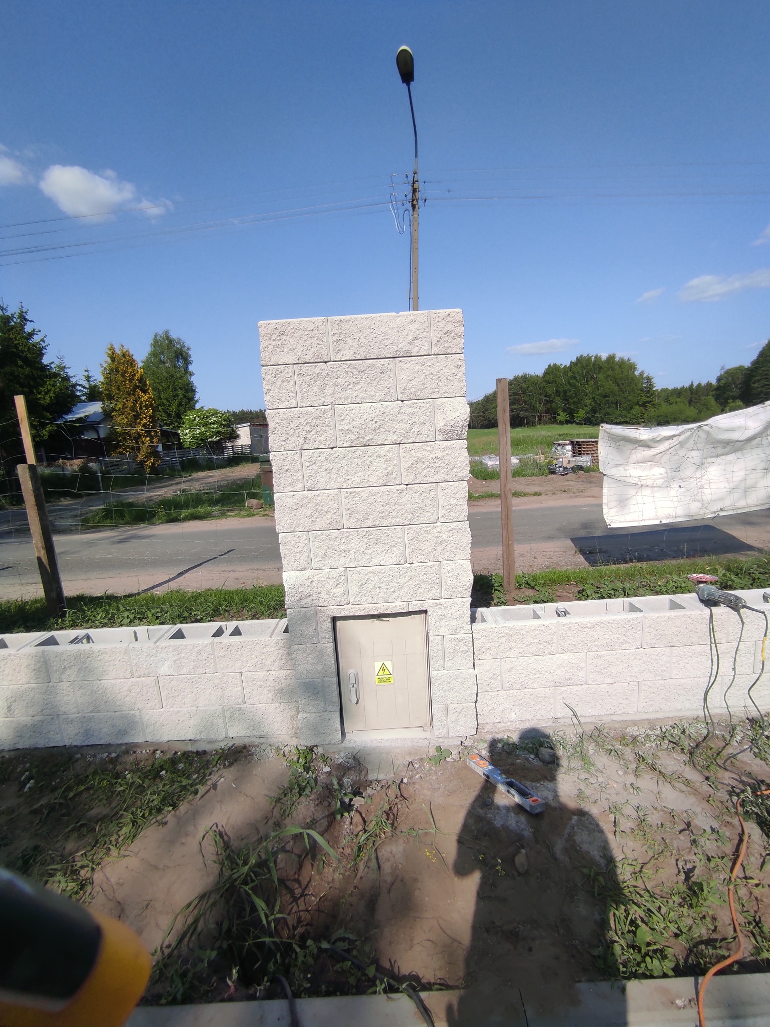 Wznoszona z szarego bloczka ogrodzeniowa ściana z wbudowaną skrzynką elektryczną, widoczny cień osoby oraz narzędzia budowlane.