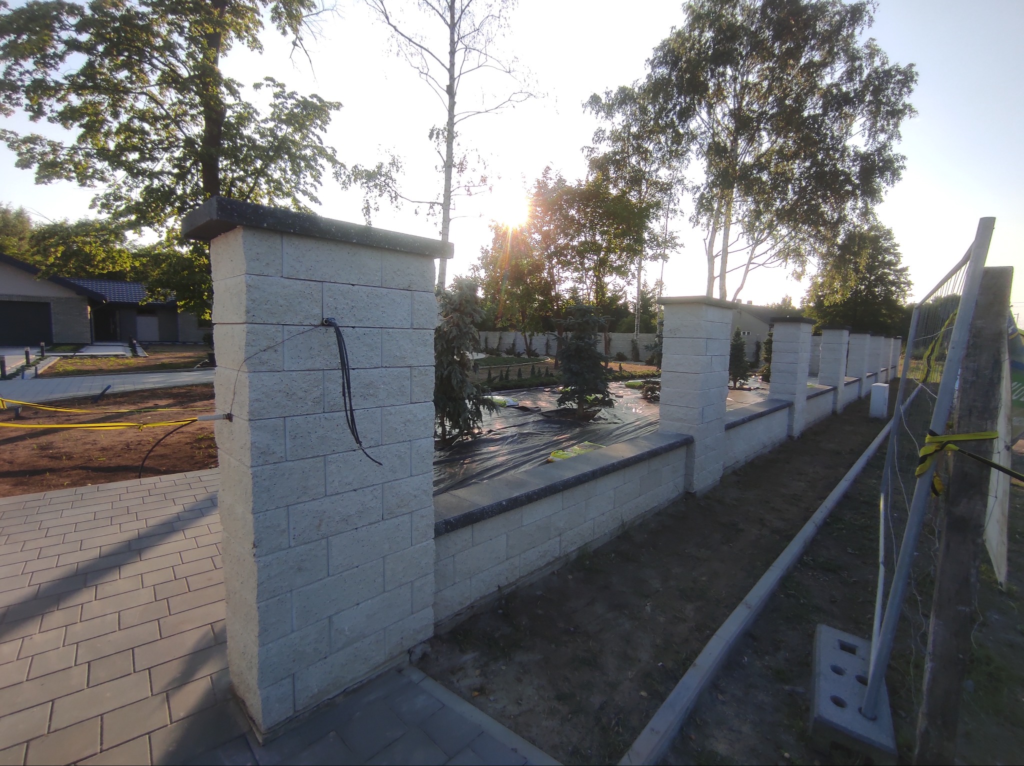 Nowo wybudowany, niski mur z jasnych, betonowych bloczków z ciemnym wykończeniem, otaczający teren z młodymi drzewkami zabezpieczonymi folią, widoczne okablowanie wystające ze słupka.