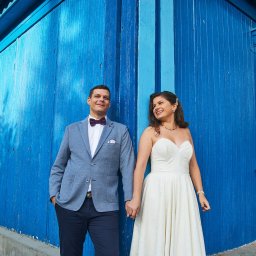 Halina Jasińska - Para młoda trzymająca się za ręce na tle błękitnej, drewnianej ściany budynku. Pan młody w marynarce i muszce, panna młoda w białej sukni.