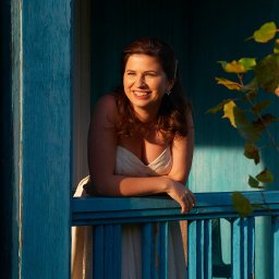 Halina Jasińska - Uśmiechnięta kobieta w białej sukience opiera się o błękitną balustradę balkonu, w tle widoczne liście i drewniana konstrukcja budynku w ciepłym świetle.