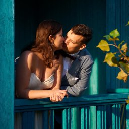 Halina Jasińska - Zakochana para w ślubnych strojach całuje się na tle turkusowej drewnianej ściany z liśćmi. Romantyczna sesja ślubna w plenerze.