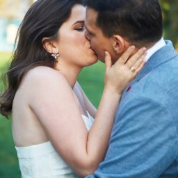 Halina Jasińska - Pocałunek pary młodej w plenerze: panna młoda w białej sukni z koronką, pan młody w błękitnej marynarce. Widoczne detale: kolczyki panny młodej, obrączka.