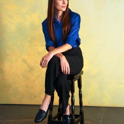 Halina Jasińska - Portret kobiety siedzącej na stołku w błękitnej koszuli i czarnych spodniach na tle żółtej ściany. Uwagę przyciągają lakierowane mokasyny.