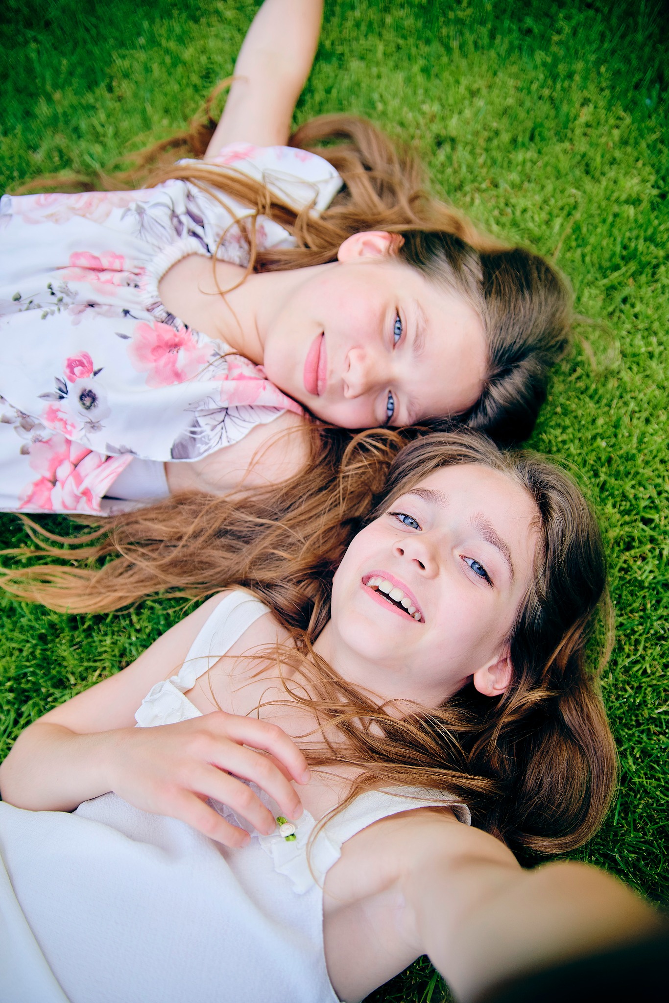 Dwie uśmiechnięte dziewczynki leżą na zielonej trawie, patrząc w obiektyw. Jedna w białej sukience, druga w sukience w kwiaty. Ujęcie z góry, naturalne światło.
