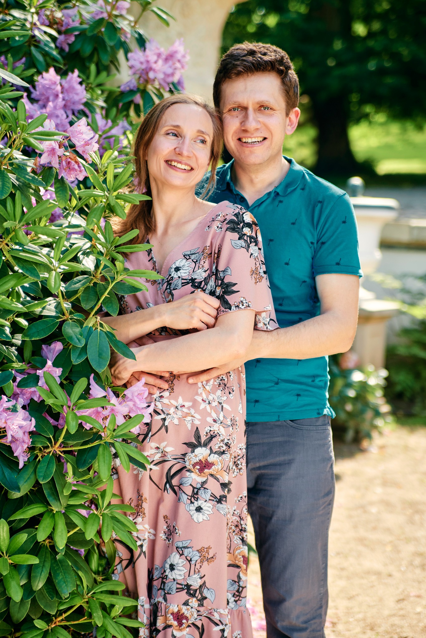 Uśmiechnięta para w objęciach, ona w kwiecistej sukience, on w turkusowej koszulce polo, pozuje przy kwitnącym krzewie w słoneczny dzień w parku.