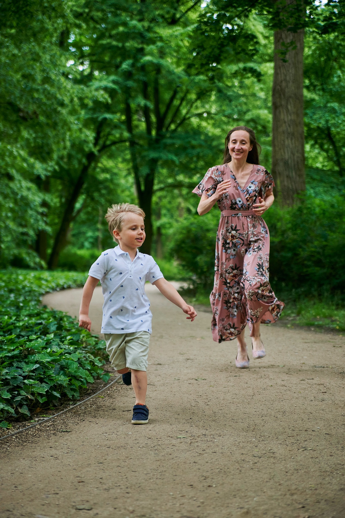 Chłopiec biegnie ścieżką w parku, za nim uśmiechnięta kobieta w kwiecistej sukience. Naturalne światło, zieleń drzew w tle. Radosna atmosfera.