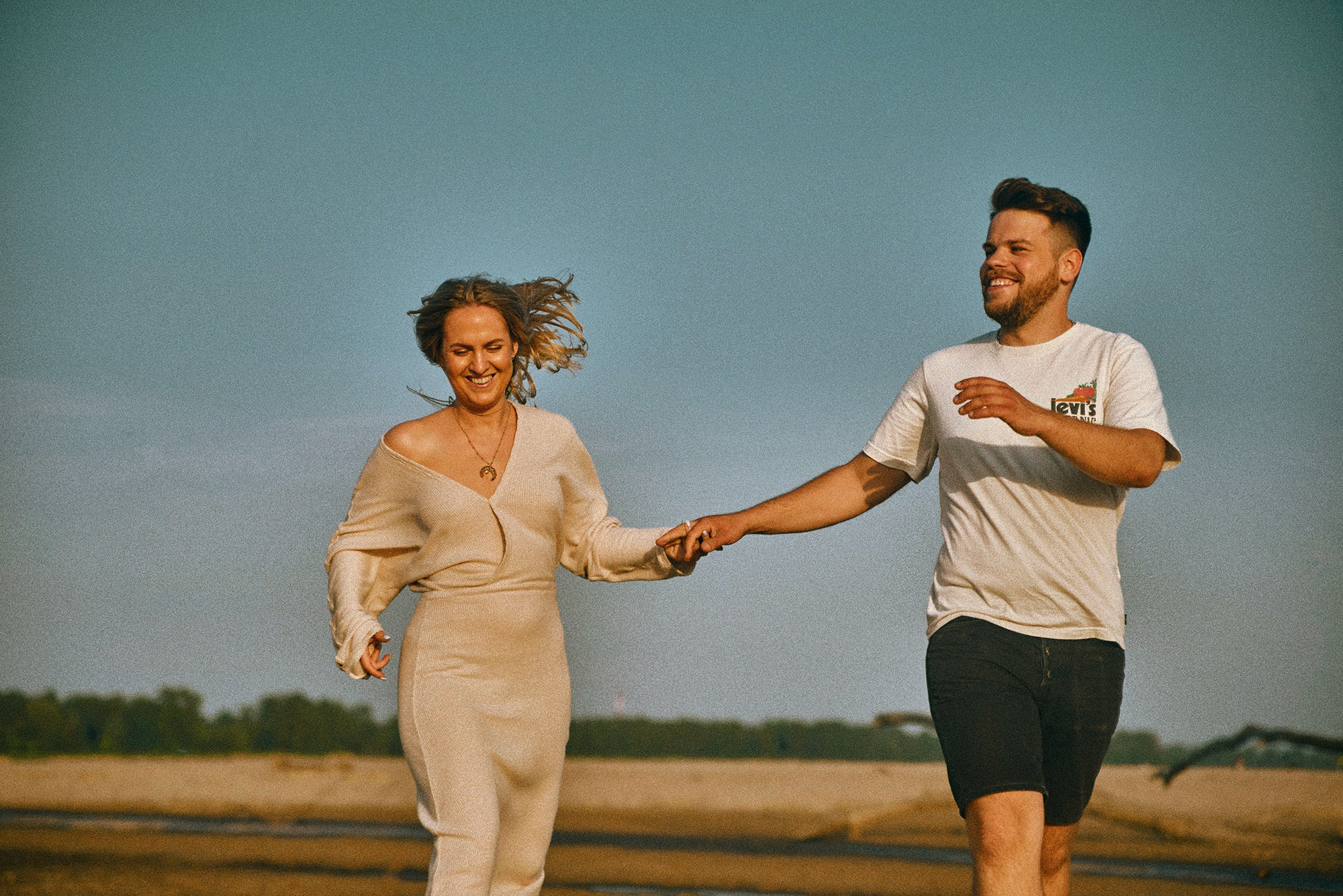 Uśmiechnięta para biegnie trzymając się za ręce na tle błękitnego nieba i piaszczystego krajobrazu. Kobieta w beżowej sukience, mężczyzna w białej koszulce i czarnych spodenkach.