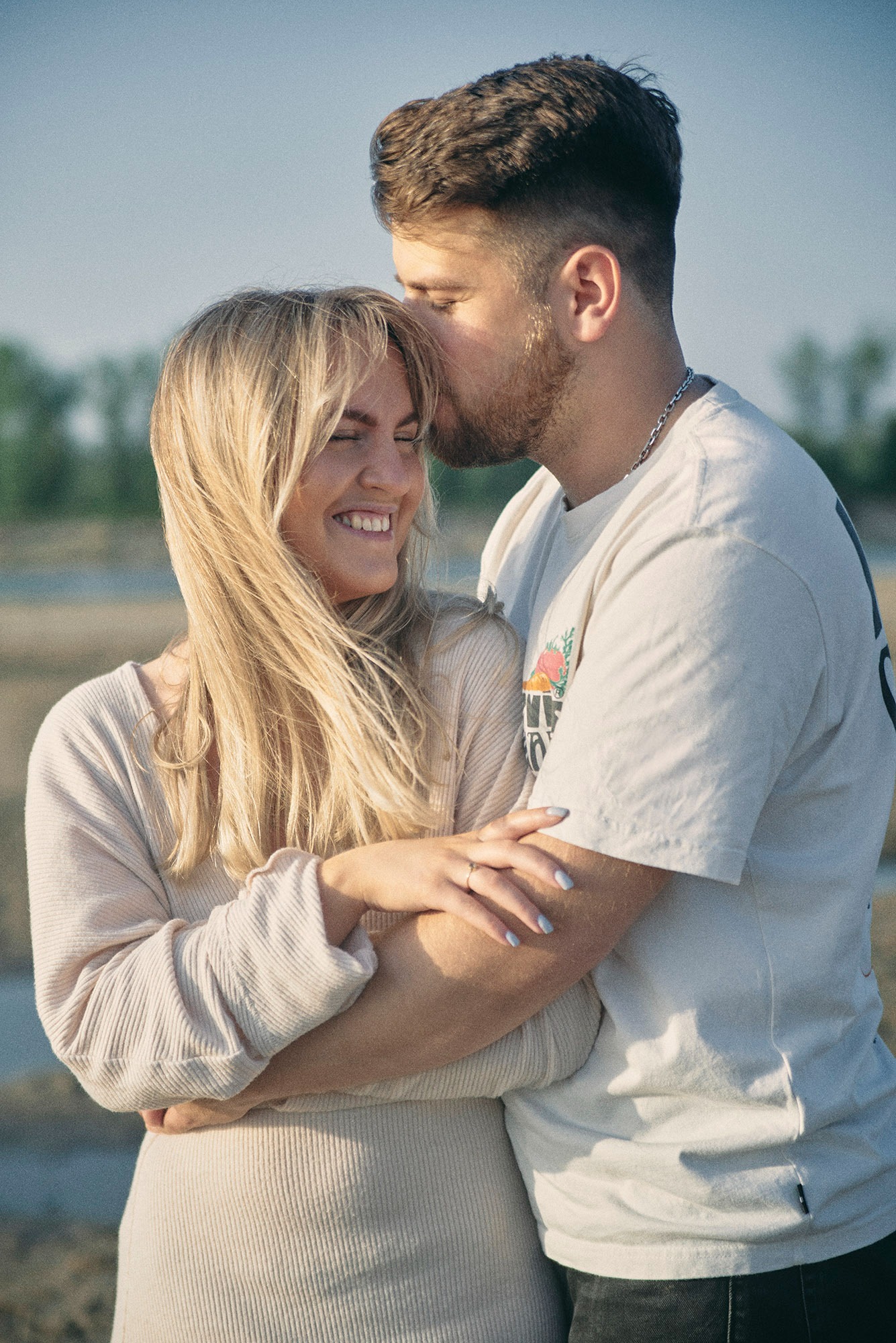 Uśmiechnięta blondynka obejmowana przez mężczyznę, który całuje ją w czoło na tle łąki. Naturalne światło, swobodna atmosfera.