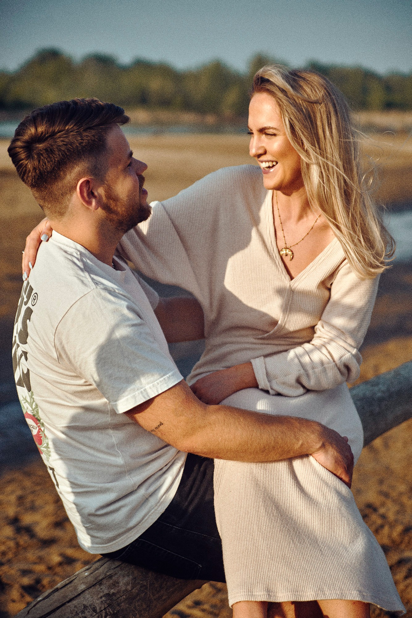Uśmiechnięta para siedzi na pniu drzewa na tle plaży. Kobieta w beżowej sukience obejmuje mężczyznę w białym T-shircie z tatuażem na ramieniu.