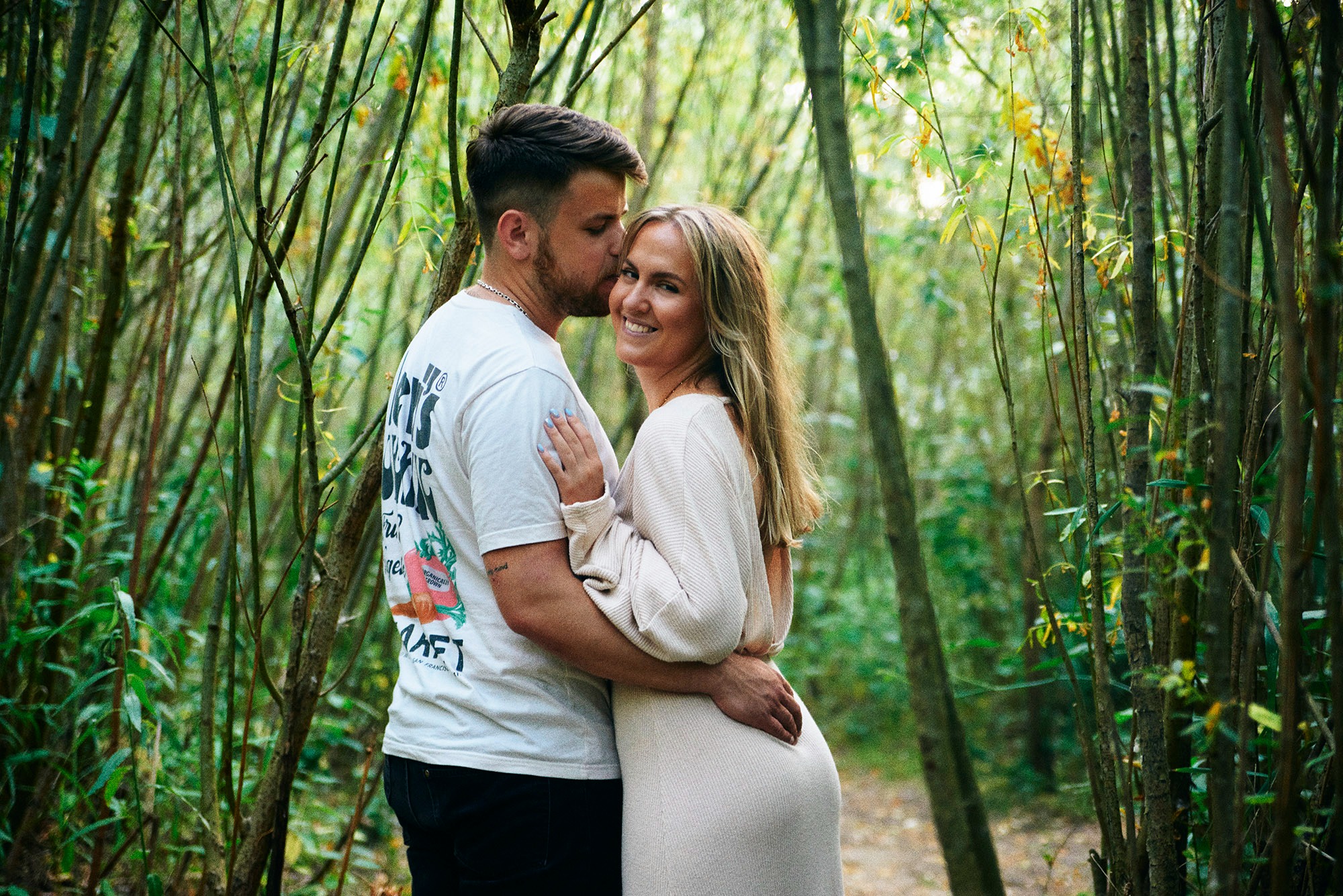 Zakochana para w objęciach na tle leśnej scenerii. Mężczyzna całuje kobietę w czoło, a ona uśmiecha się do kamery. Naturalne światło, ciepłe barwy, romantyczna atmosfera.