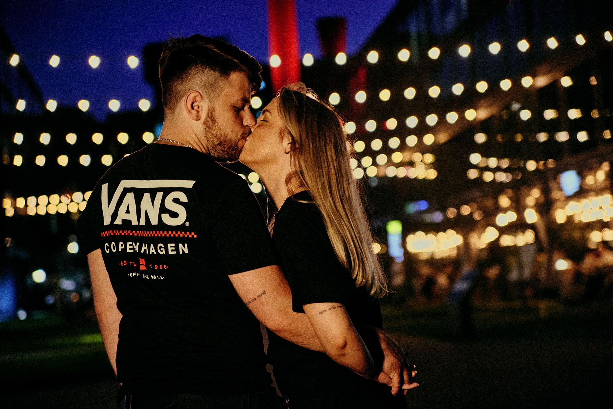 Zakochana para całuje się na tle miejskich świateł. Mężczyzna w koszulce Vans, kobieta z blond włosami, oboje z tatuażami na ramionach. Ujęcie z bliska, nocna sceneria.