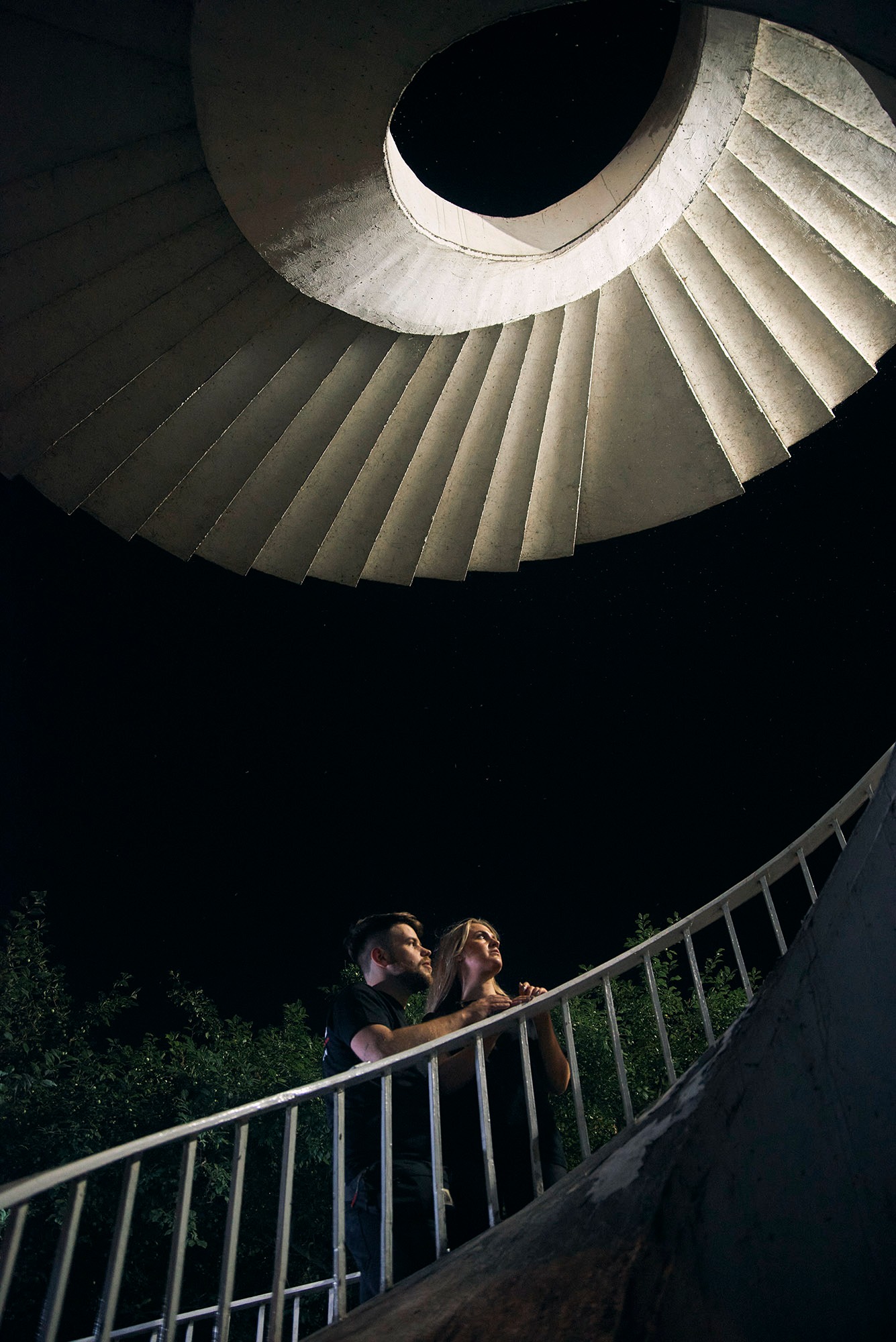Para na tle spiralnych schodów pod gołym niebem, nocą. Mężczyzna obejmuje kobietę, oboje patrzą w górę. Nietypowe ujęcie z dołu, widoczne gwiazdy.