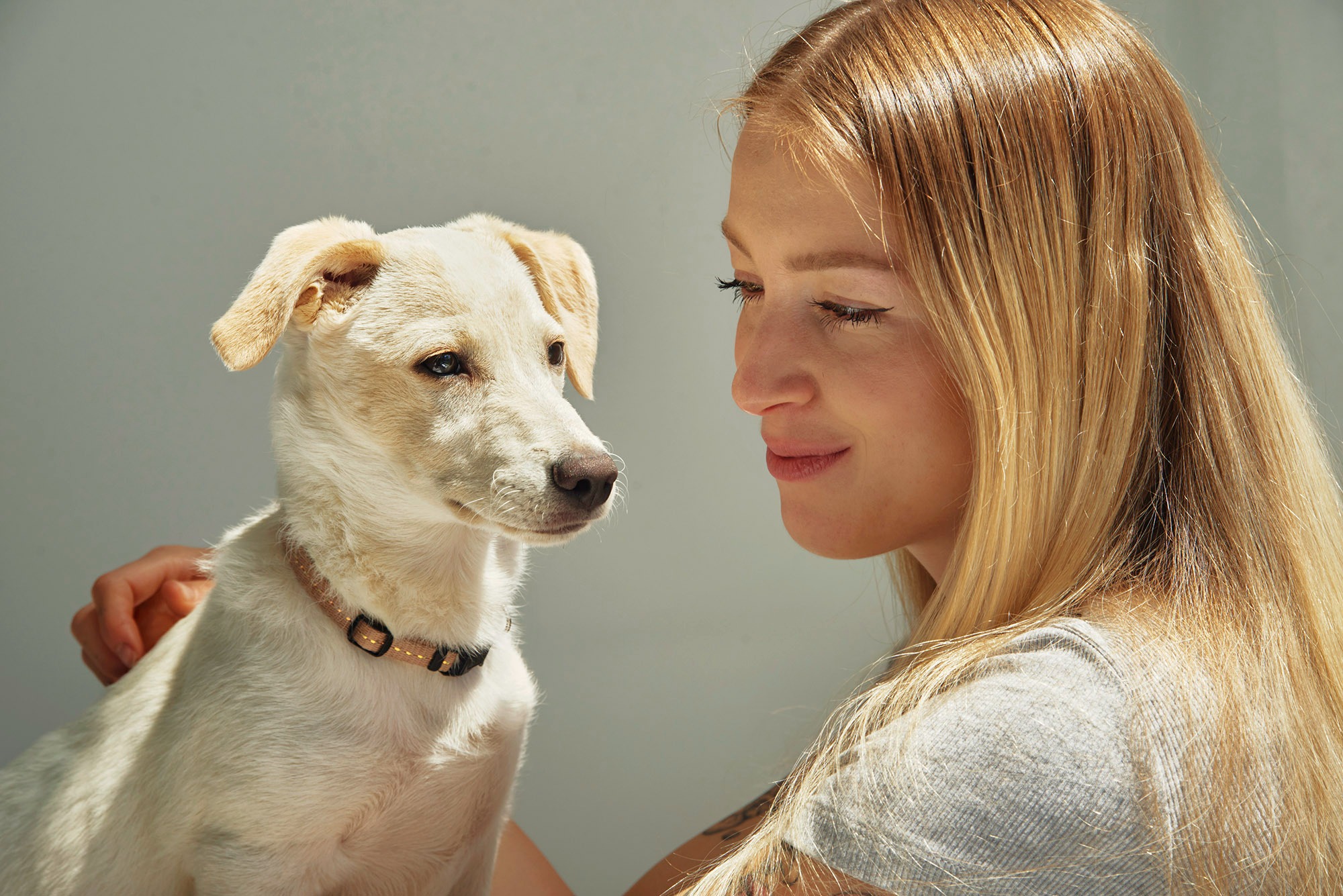Portret młodej kobiety z długimi blond włosami, spoglądającej na jasnego szczeniaka w obroży. Delikatne światło podkreśla ich bliskość.