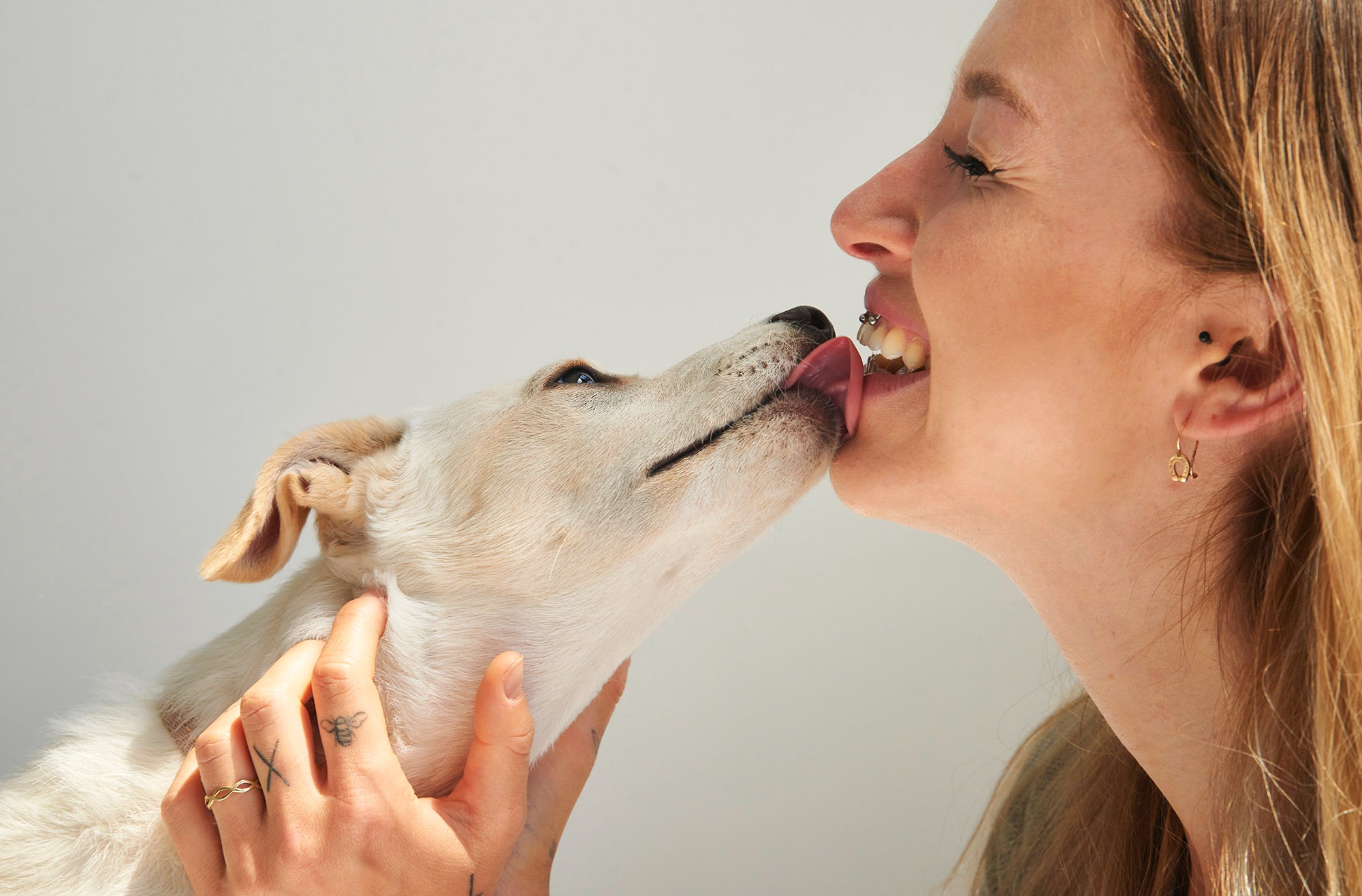 Kobieta z piercingiem w wardze, profil, całowana przez psa rasy mieszanej z jasną sierścią; widoczne tatuaże na dłoni kobiety i złote kolczyki w kształcie podkowy.