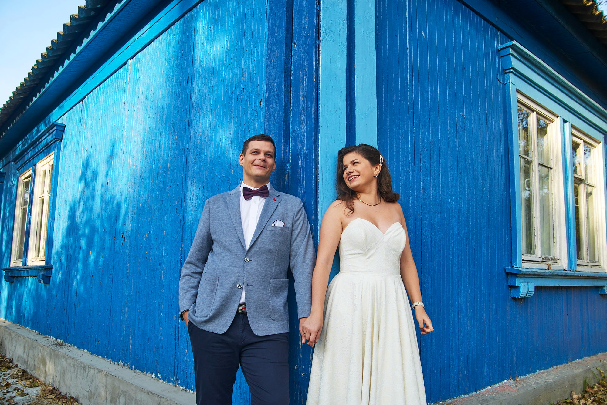 Para młoda trzymająca się za ręce na tle błękitnej, drewnianej ściany budynku. Pan młody w marynarce i muszce, panna młoda w białej sukni.