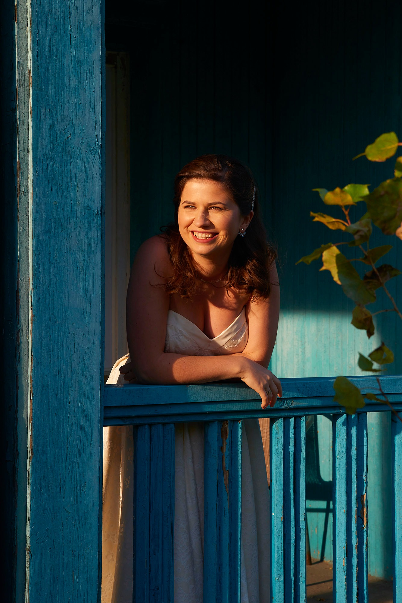 Uśmiechnięta kobieta w białej sukience opiera się o błękitną balustradę balkonu, w tle widoczne liście i drewniana konstrukcja budynku w ciepłym świetle.