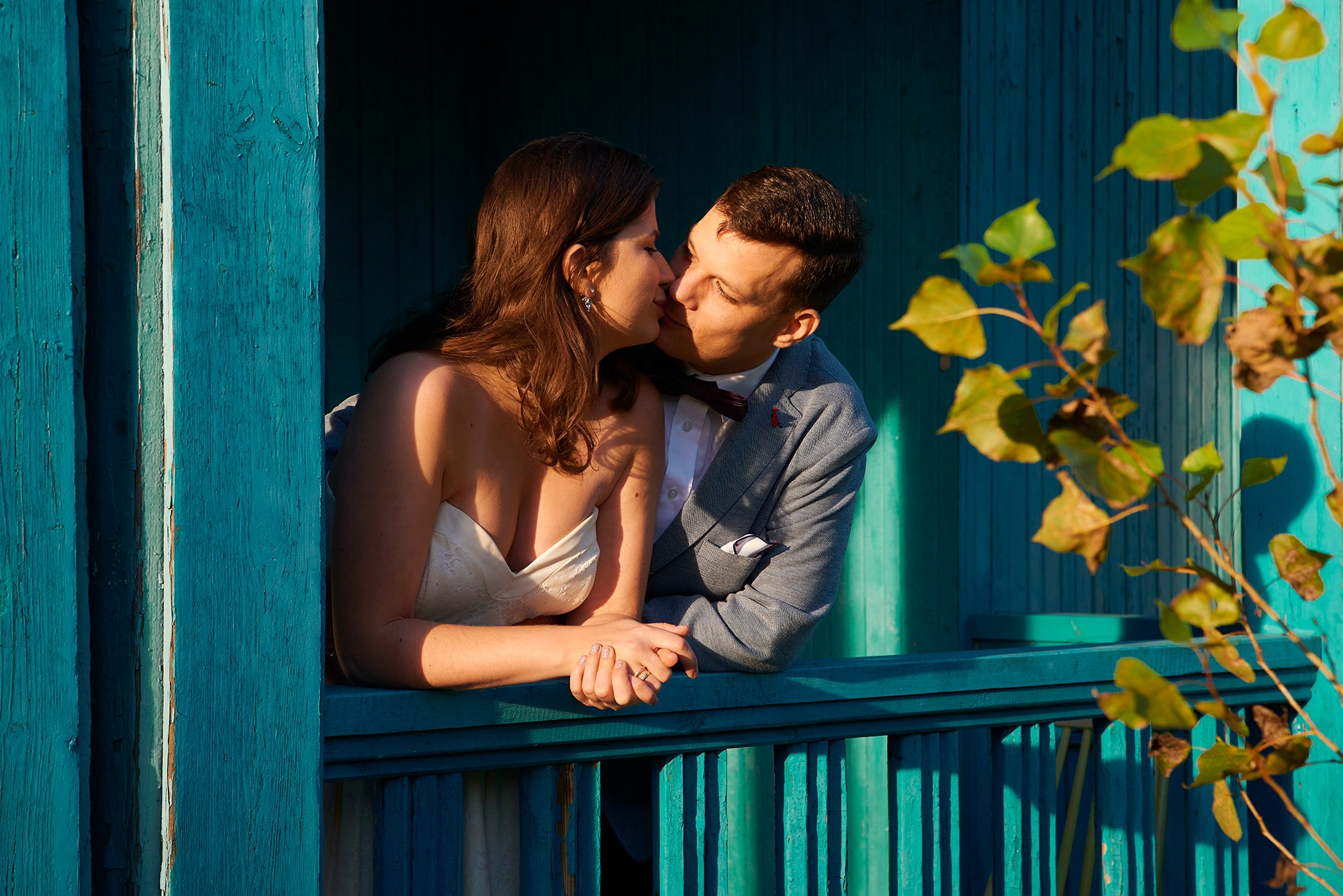 Zakochana para w ślubnych strojach całuje się na tle turkusowej drewnianej ściany z liśćmi. Romantyczna sesja ślubna w plenerze.