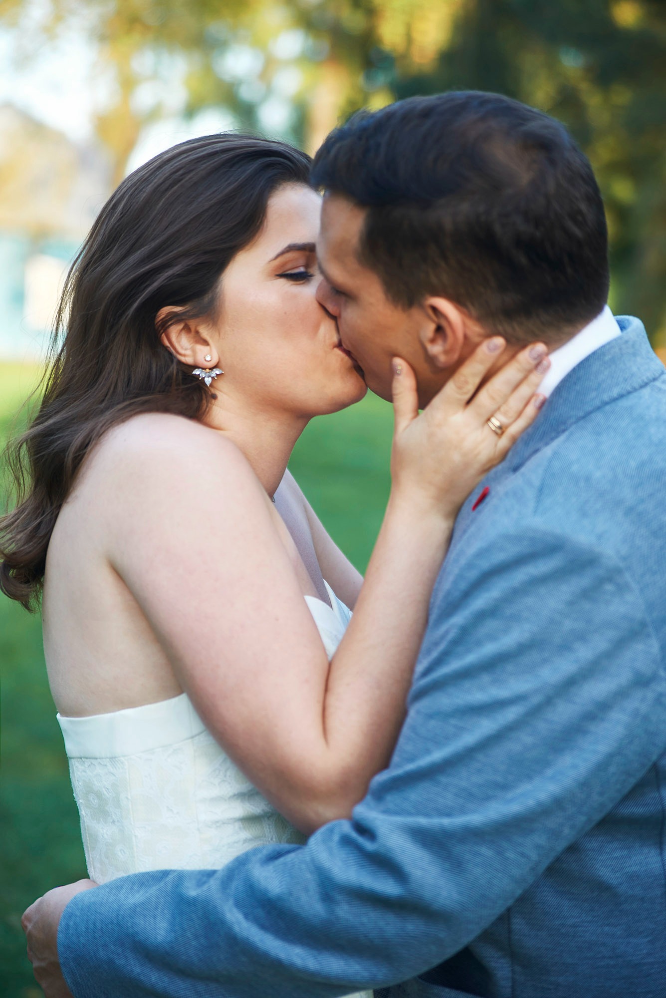 Pocałunek pary młodej w plenerze: panna młoda w białej sukni z koronką, pan młody w błękitnej marynarce. Widoczne detale: kolczyki panny młodej, obrączka.