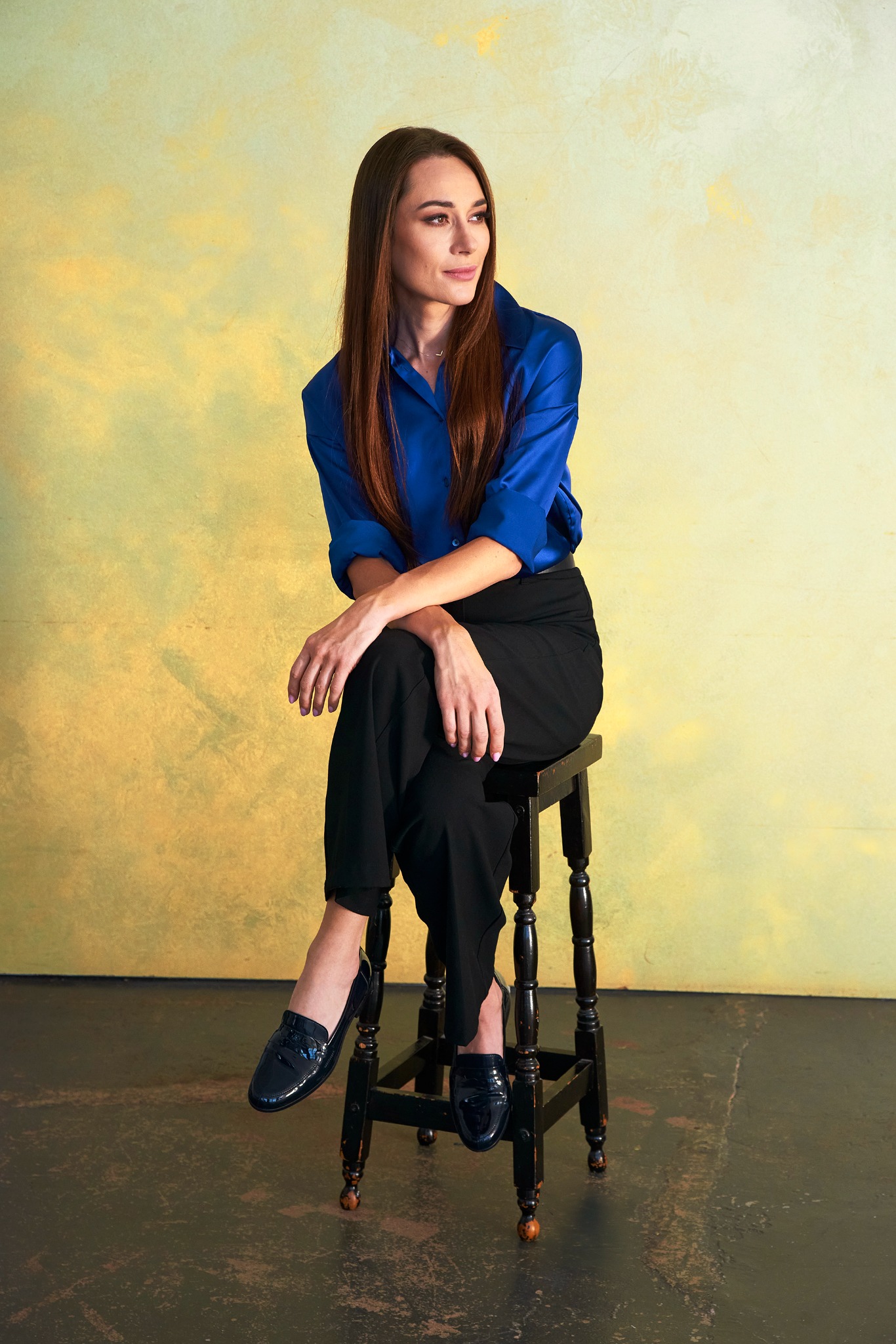 Portret kobiety siedzącej na stołku w błękitnej koszuli i czarnych spodniach na tle żółtej ściany. Uwagę przyciągają lakierowane mokasyny.