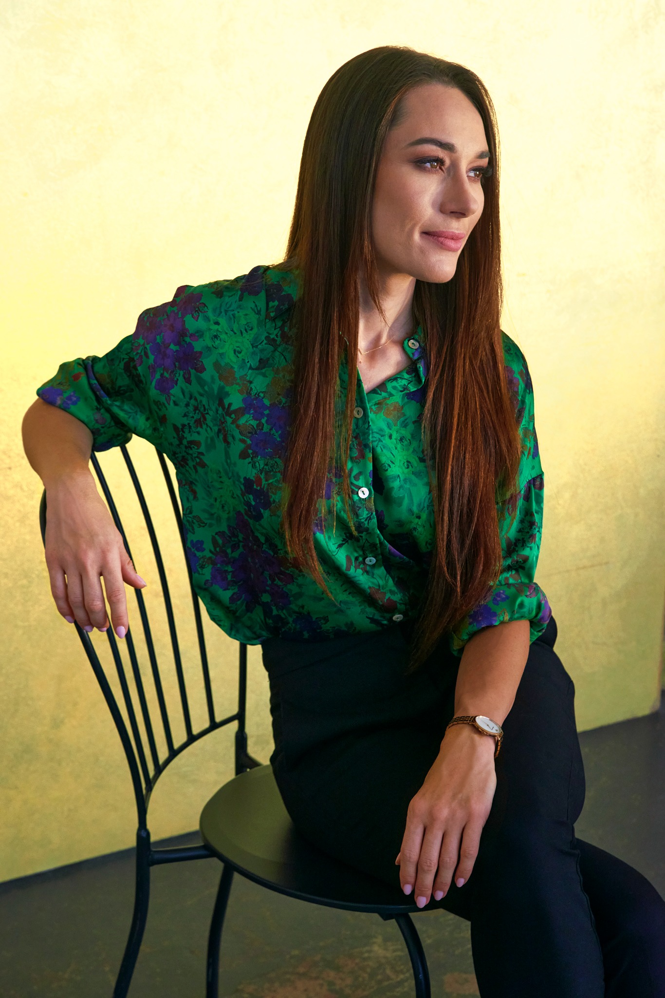 Portret kobiety w zielonej koszuli w kwiaty, siedzącej na czarnym krześle na tle żółtej ściany. Delikatny uśmiech, długie włosy i subtelny makijaż.