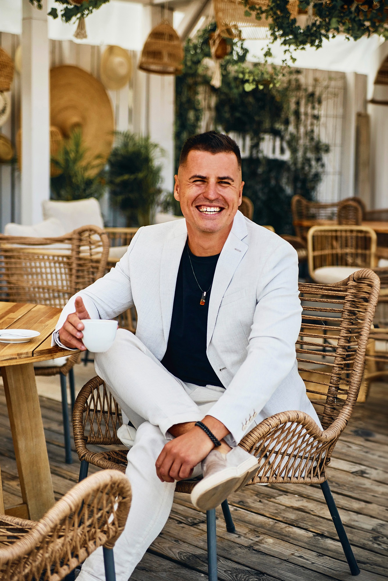 Uśmiechnięty mężczyzna w białym garniturze siedzi w wiklinowym fotelu z filiżanką. Jasne tło z roślinami i dekoracjami. Swobodna elegancja, ciepłe światło.