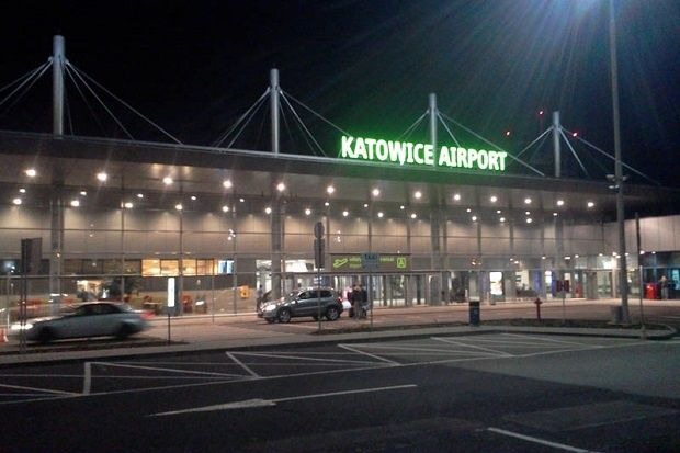 Nocny widok na oświetlony budynek lotniska Katowice-Pyrzowice z zaparkowanymi samochodami na pierwszym planie.