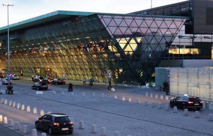 Nowoczesny budynek lotniska z charakterystyczną szklaną fasadą w kształcie rombów; przed budynkiem zaparkowane samochody i osoby oczekujące na transport.