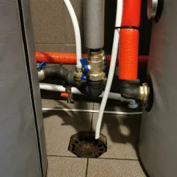 Fragment instalacji z rurami w izolacji termicznej, zaworami z niebieskimi i pomarańczowymi uchwytami oraz elementem odpływowym na szarej, kafelkowej podłodze, widoczne elementy zbiornika.