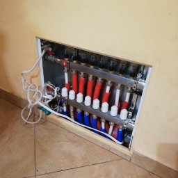 Hitmen Construction - Rozdzielacz ogrzewania podłogowego z czerwonymi i niebieskimi rurami, widoczny w otworze w ścianie, z przewodami wystającymi na zewnątrz.