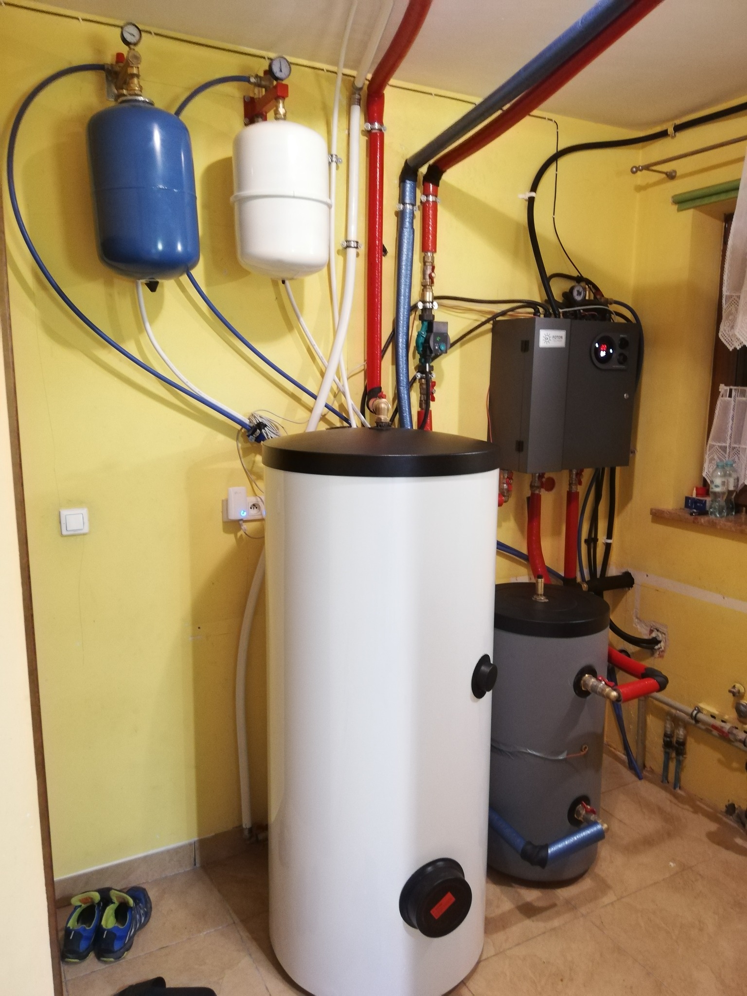 Biały i szary zbiornik buforowy z podłączonymi rurami w kotłowni o żółtych ścianach, widoczne elementy instalacji i osprzęt.