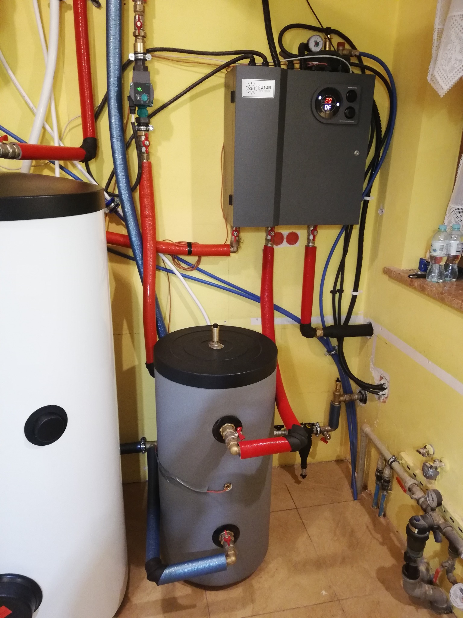 Dwa zbiorniki akumulacyjne wody, biały i szary, podłączone do szarej jednostki sterującej z wyświetlaczem temperatury 20 stopni Celsjusza, z widocznymi rurami w izolacji czerwonej i niebieskiej...