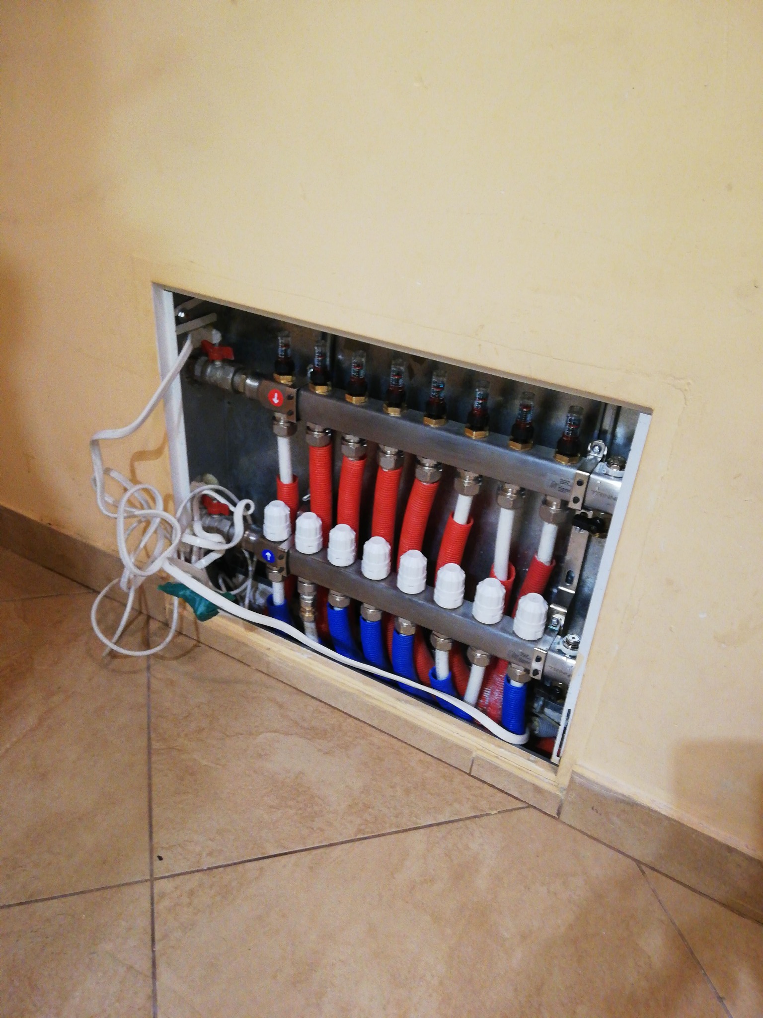 Rozdzielacz ogrzewania podłogowego z czerwonymi i niebieskimi rurami, widoczny w otworze w ścianie, z przewodami wystającymi na zewnątrz.