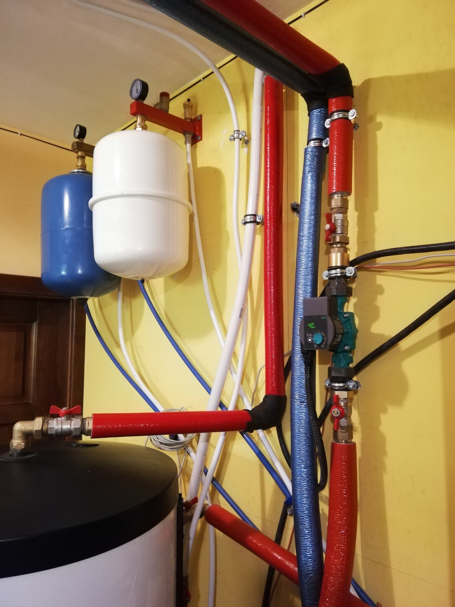 Złożona instalacja hydrauliczna z pompą obiegową Wilo, zbiornikami wyrównawczymi (niebieski i biały) oraz rurami w izolacji czerwonej i niebieskiej, zamontowana na żółtej ścianie.