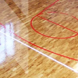 Parkietowa podłoga hali sportowej z wyraźnymi czerwonymi liniami boiska i odblaskami światła, ukazująca odnowioną powierzchnię.