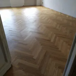 Nowo ułożony parkiet w jodełkę w jasnym, pustym pokoju z dwoma oknami i białymi ścianami, widok zza otwartych drzwi.