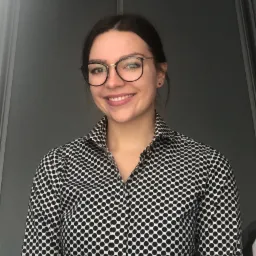 Uśmiechnięta kobieta w okularach i koszuli w czarne kropki na szarym tle.