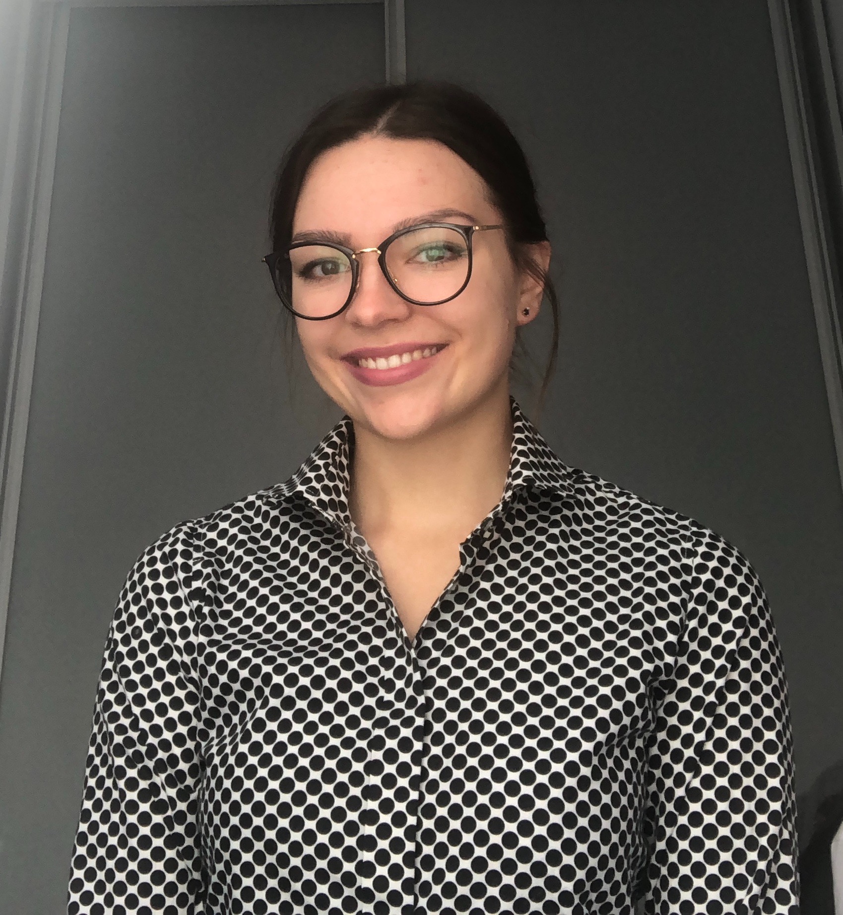 Uśmiechnięta kobieta w okularach i koszuli w czarne kropki na szarym tle.