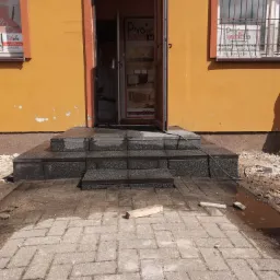 Nowe, granitowe schody wejściowe do budynku z pomarańczową elewacją, widoczne ślady prac wykończeniowych na chodniku z kostki brukowej.