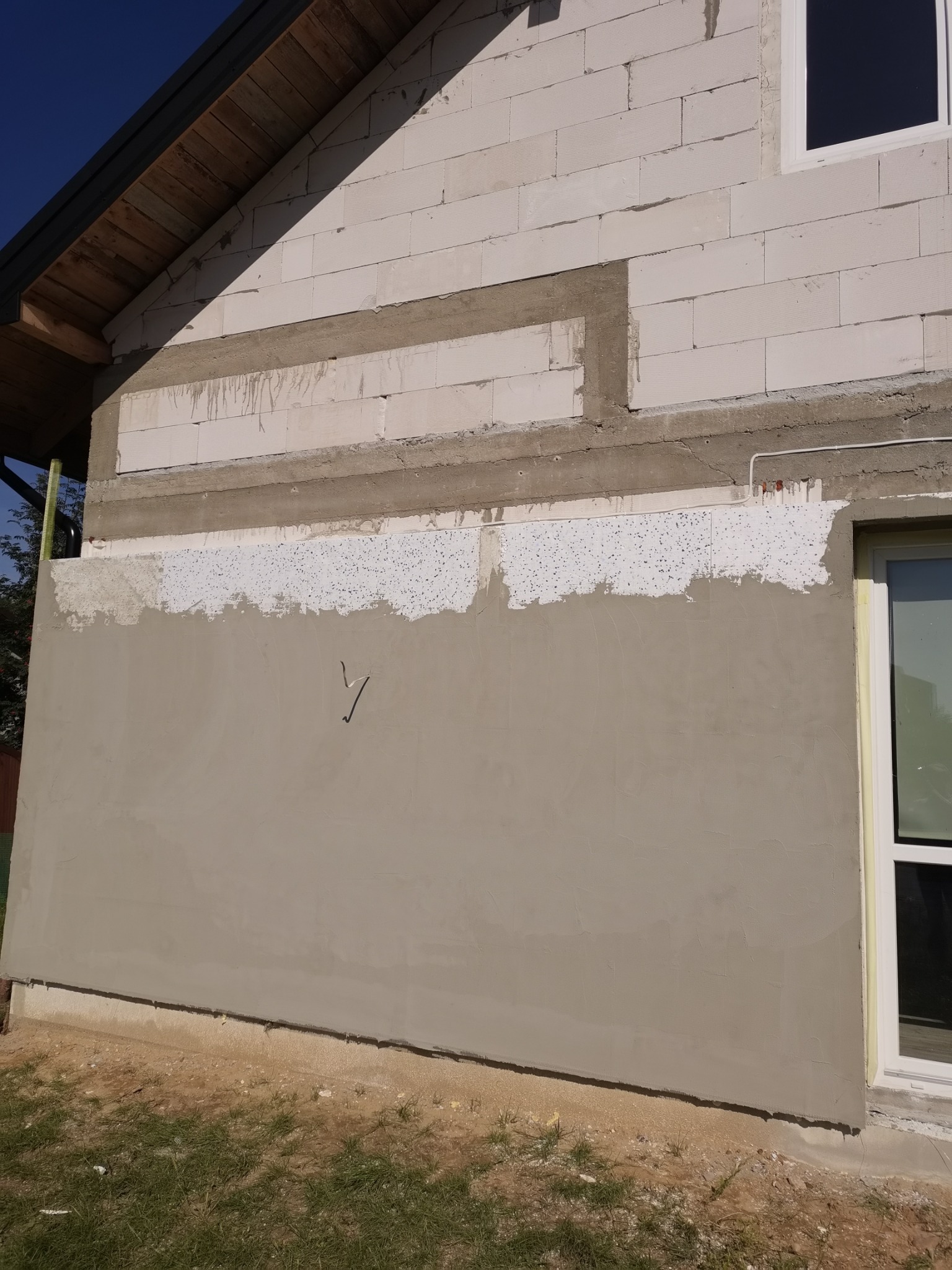 Ściana zewnętrzna budynku w trakcie prac wykończeniowych: fragment ocieplenia z widoczną strukturą styropianu, poniżej nałożona warstwa świeżej, jasnoszarej wyprawy tynkarskiej, z widocznymi...