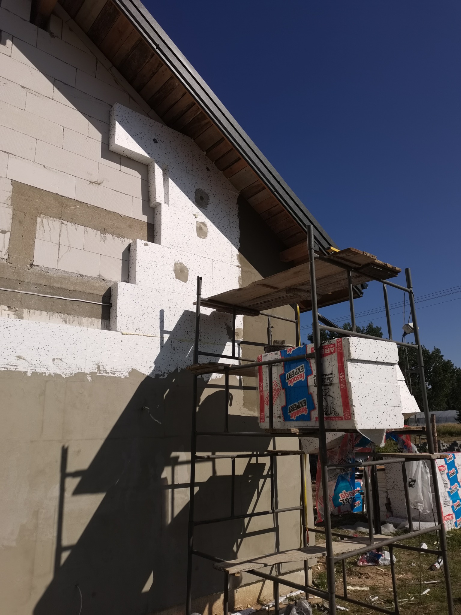 Ocieplanie ściany zewnętrznej budynku z białej cegły bloczkowej, widoczne fragmenty przyklejonego styropianu i rusztowanie z paczkami styropianu marki Fasada Expert.