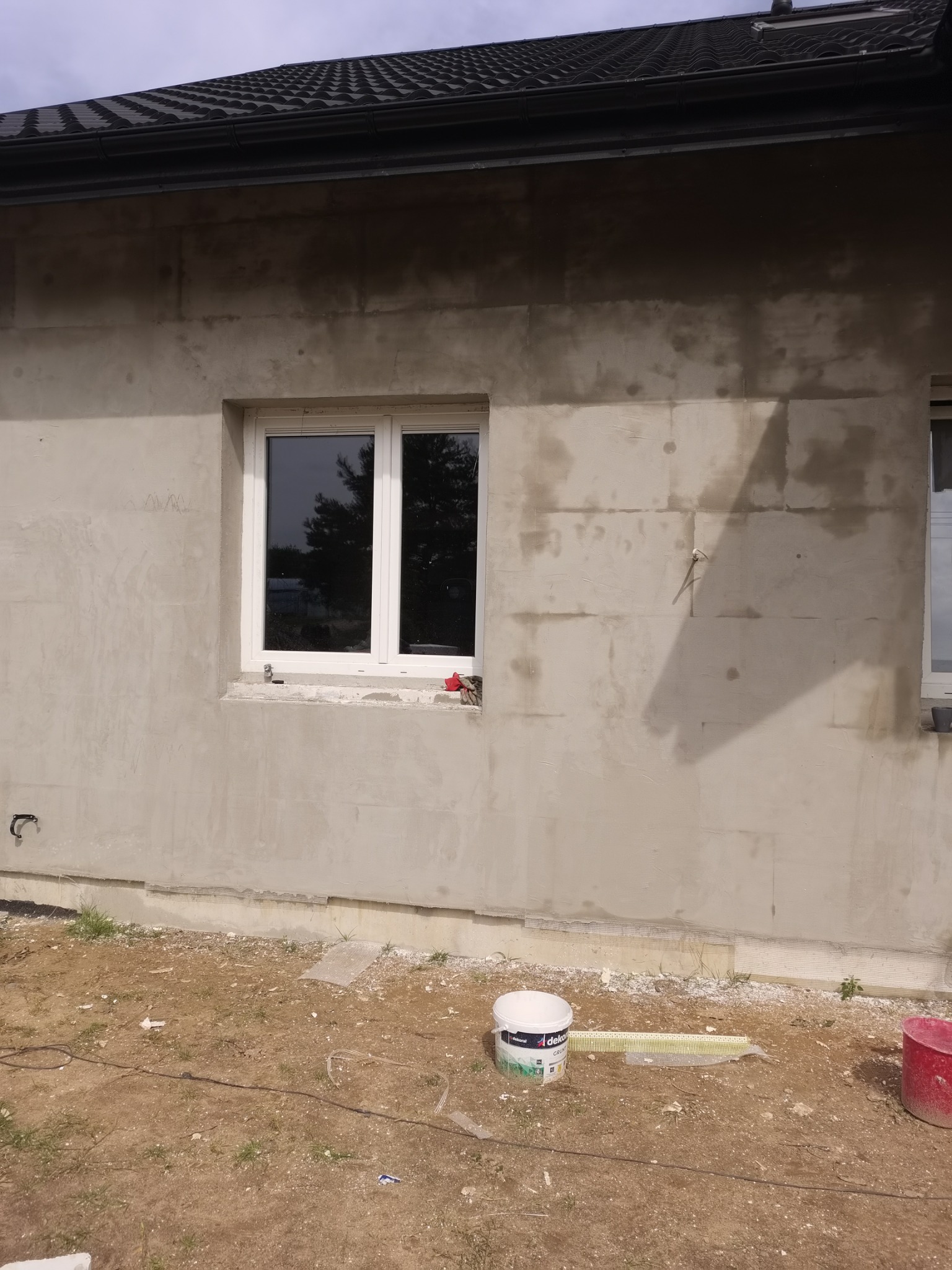 Surowa elewacja domu z oknem przed nałożeniem warstwy wykończeniowej, widoczne ślady po szpachlowaniu, wiadro z masą szpachlową Dekoral i miarka leżące na ziemi.