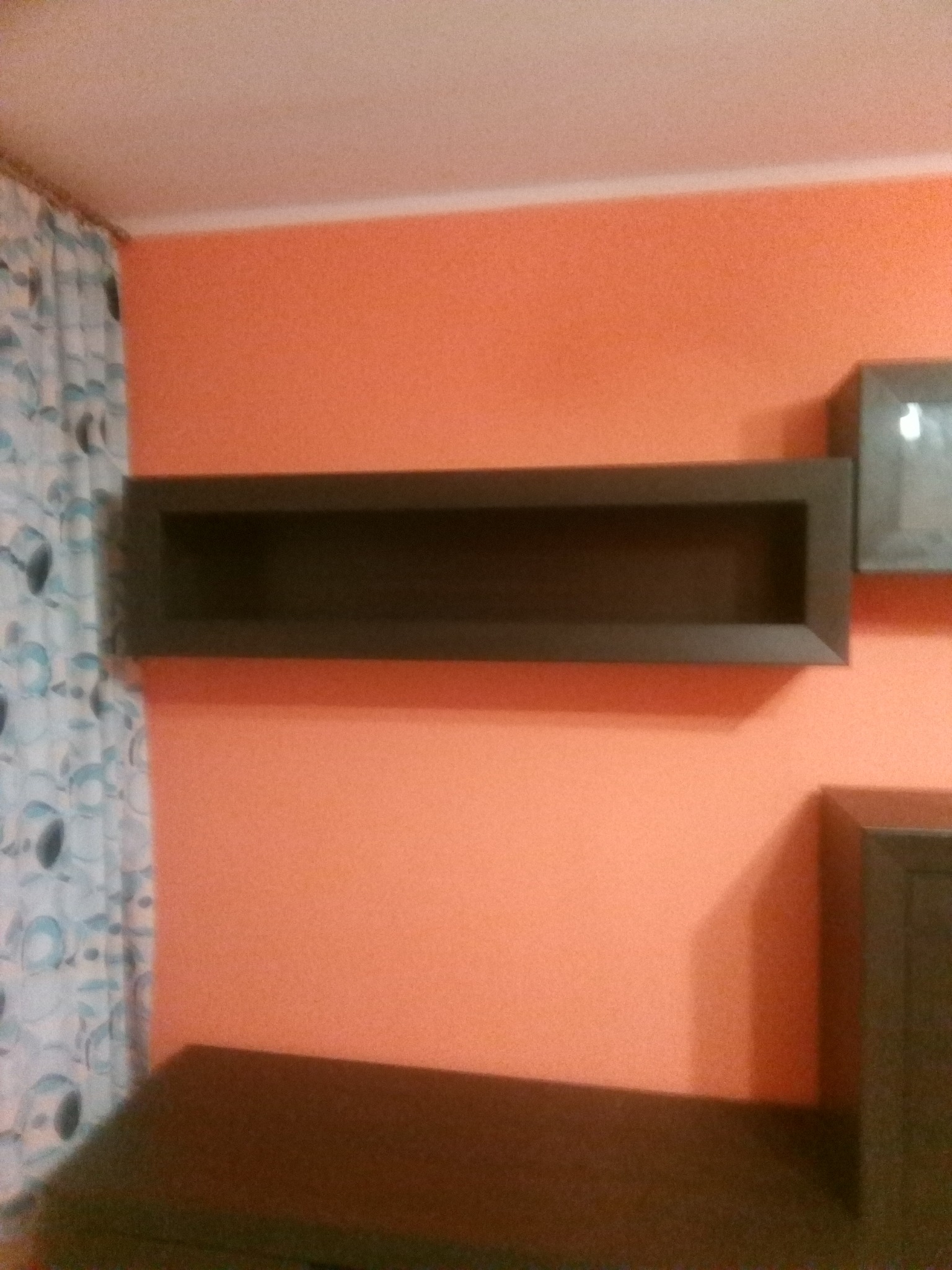 Pomalowana na pomarańczowo ściana z zamontowanymi półkami w kolorze ciemnego brązu, zasłonięta częściowo zasłoną z niebieskim wzorem.
