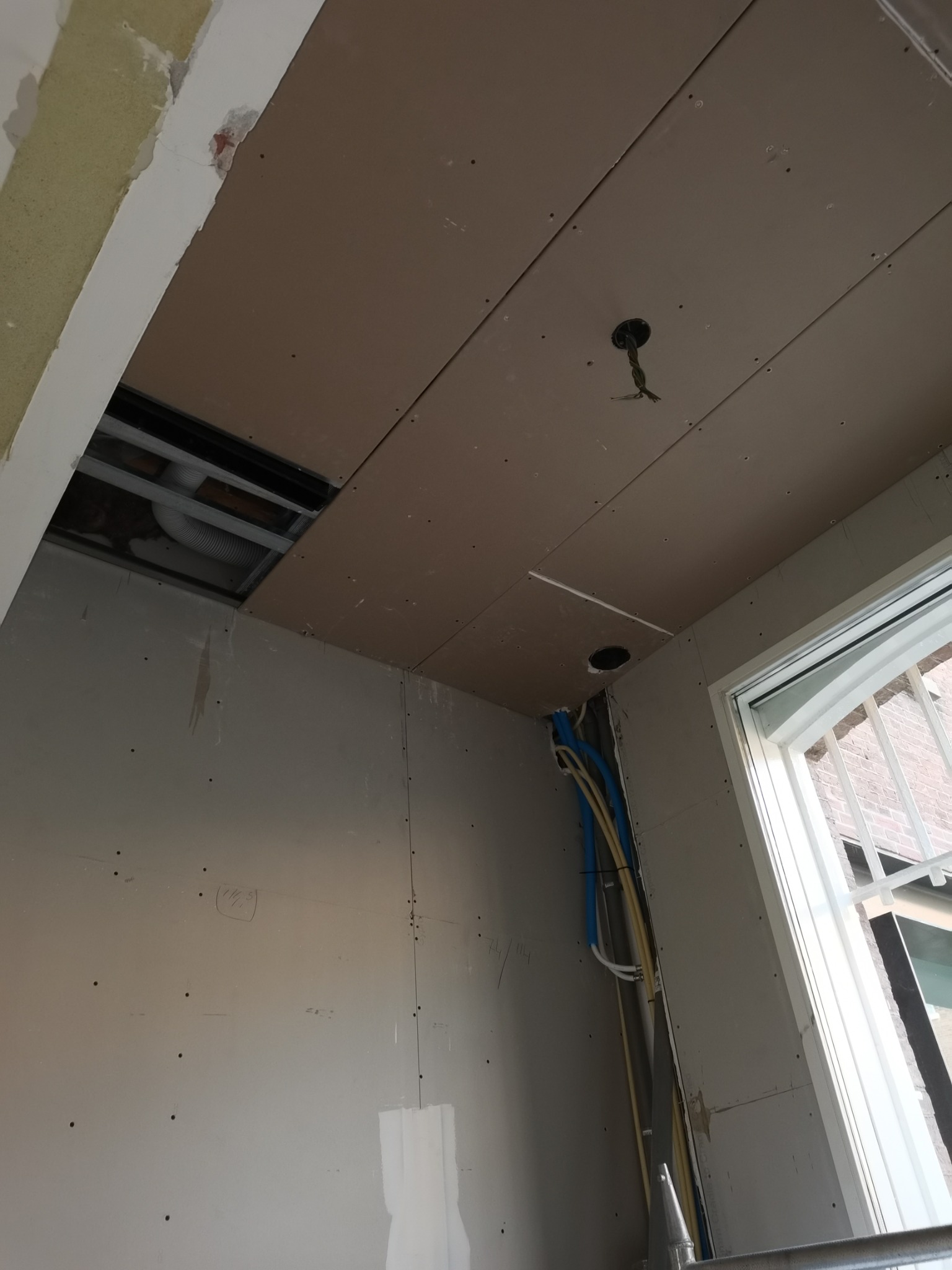 Wnętrze w trakcie prac wykończeniowych: zamontowane płyty kartonowo-gipsowe na suficie i ścianach, widoczne otwory na oświetlenie i instalacje elektryczne, fragment okna z kratą w tle.