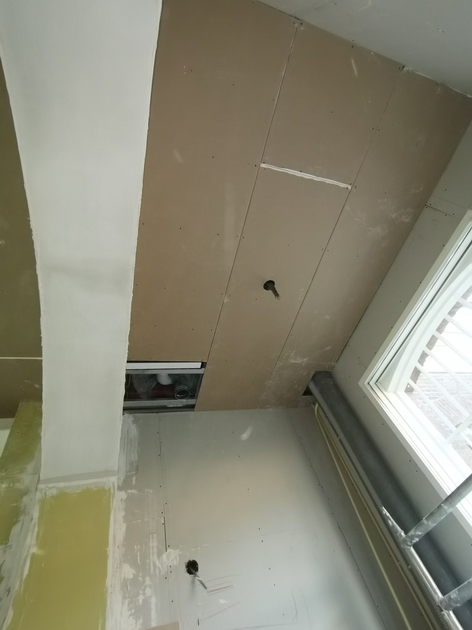 Sufit z płyt kartonowo-gipsowych w trakcie montażu, widoczne łączenia płyt, otwór na instalację elektryczną i fragment okna z rusztowaniem.