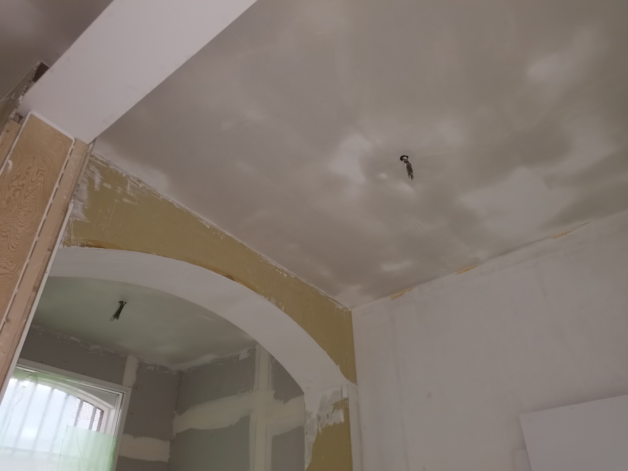 Wnętrze w trakcie remontu: sufit i ściany po szpachlowaniu, widoczne przewody elektryczne zwisające z sufitu, przygotowane do dalszych prac wykończeniowych, łukowe przejście między pomieszczeniami.