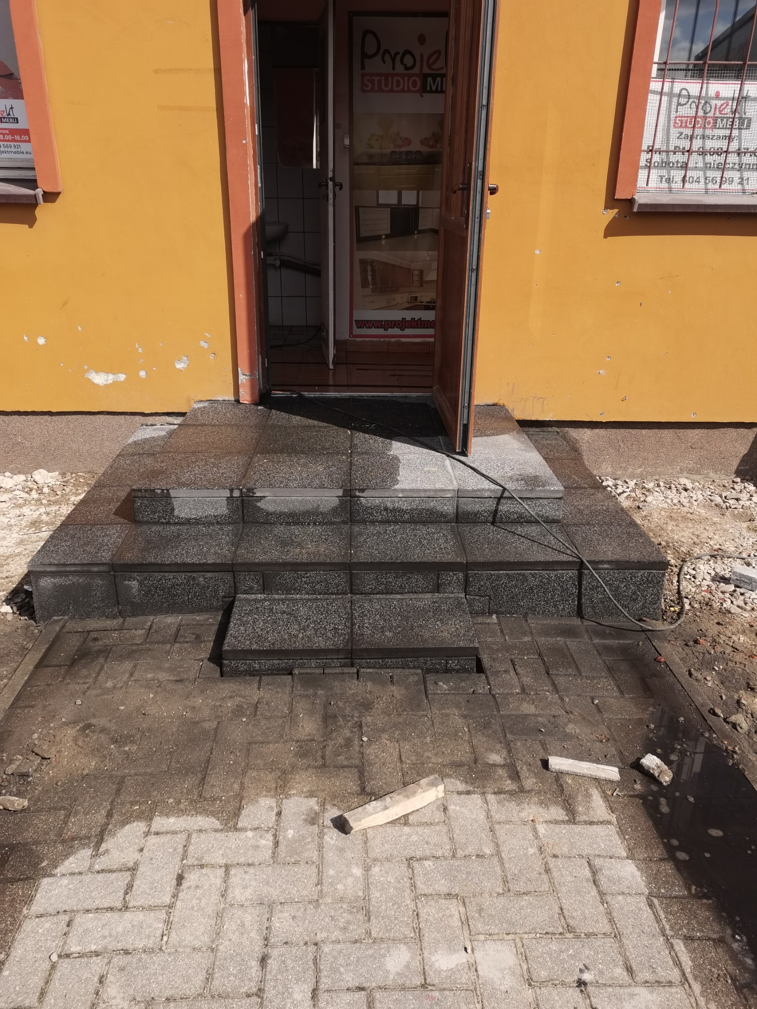 Wykonane schody wejściowe z ciemnej kostki granitowej prowadzące do budynku z żółtą elewacją i otwartymi drzwiami, widoczne fragmenty kostki brukowej na podłożu.