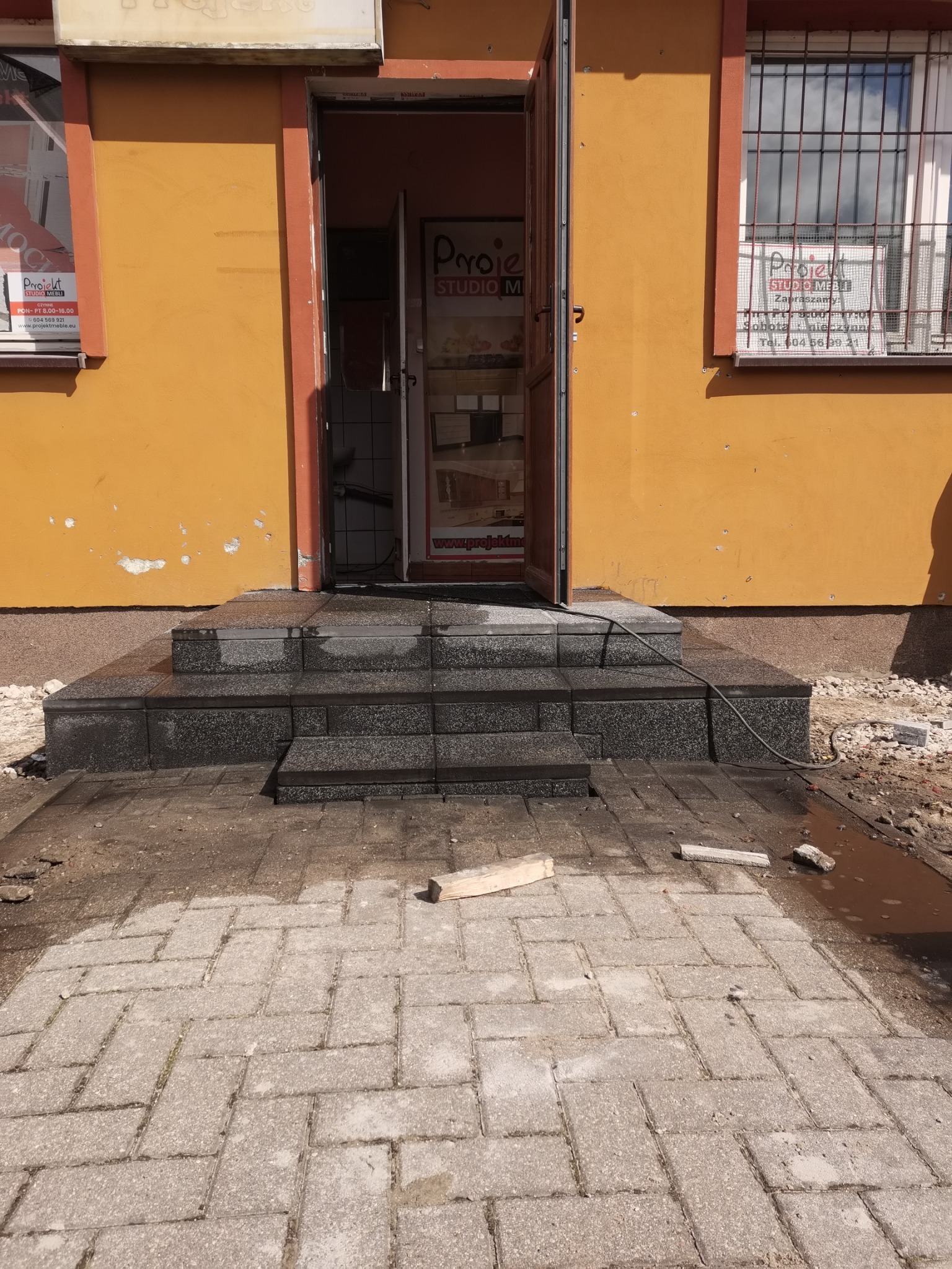 Nowe, granitowe schody wejściowe do budynku z pomarańczową elewacją, widoczne ślady prac wykończeniowych na chodniku z kostki brukowej.